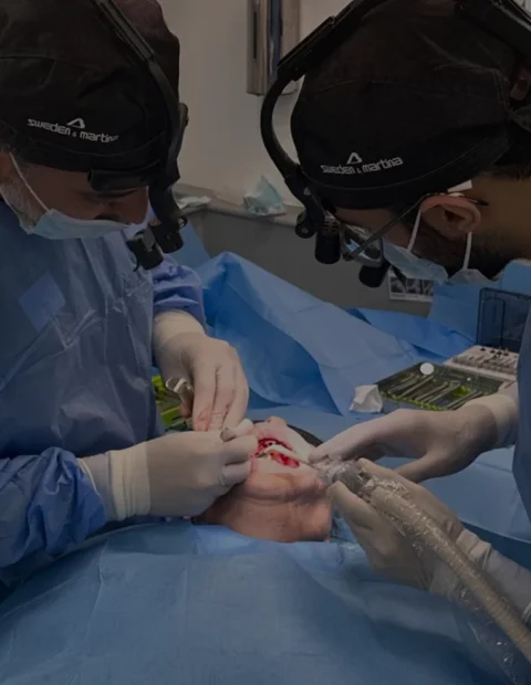 Two surgeons in scrubs and loupes performing oral surgery on a patient